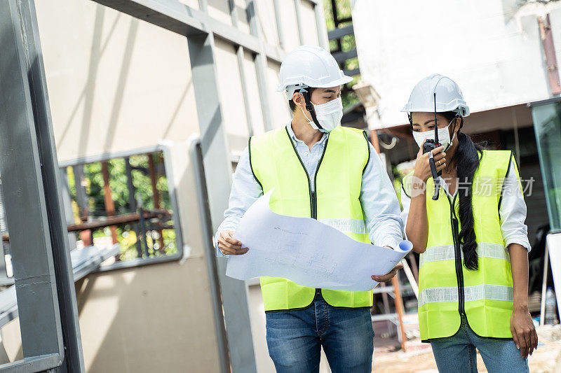 亚洲工人在建筑现场佩戴防护口罩。男和女工程师拿着图纸在施工方讨论项目。