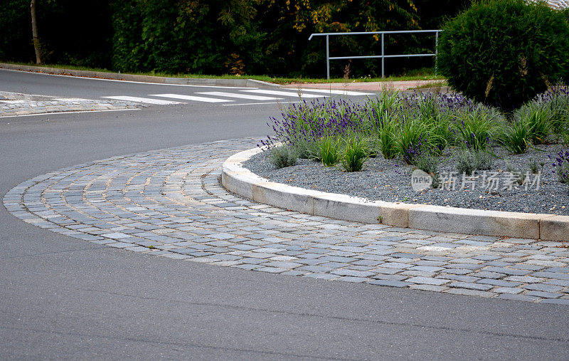 环形路铺设灰色花岗岩立方体，交通枢纽，中间有鲜花和草。针叶树呈绿色球状，开花淡紫色蓝色，砾石覆盖