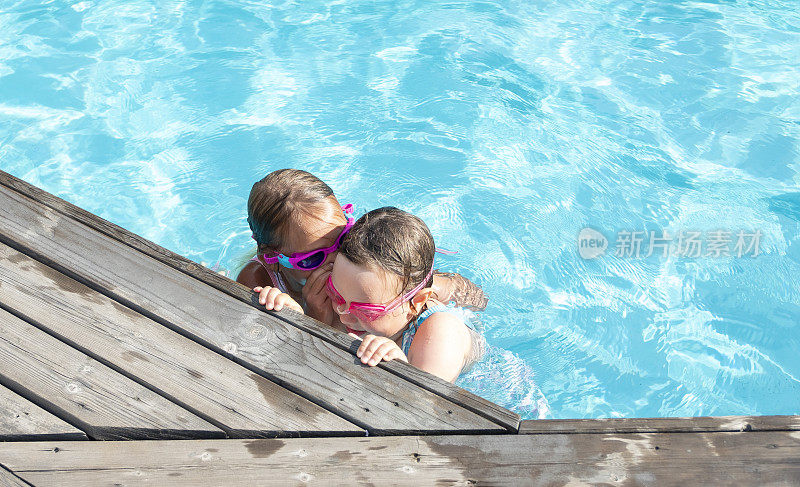 女孩，白种人的孩子，在室外游泳池游泳。两个朋友互相拥抱，低声耳语。