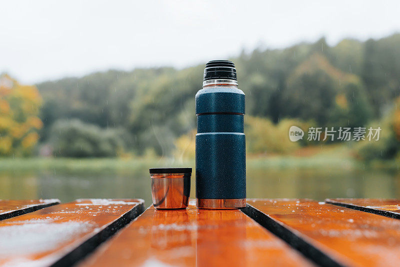 保温瓶和铝制马克杯，里面装着热饮和户外上升的蒸汽。在阴雨、寒冷、多云的天气里，放在湖边和森林附近的木桌上的野营保温瓶
