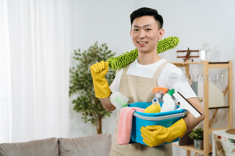 微笑的年轻亚洲韩国人与一桶洗涤剂的手在橡胶保护手套持有。保洁家政服务理念。帅哥面对镜头自信的微笑享受家务