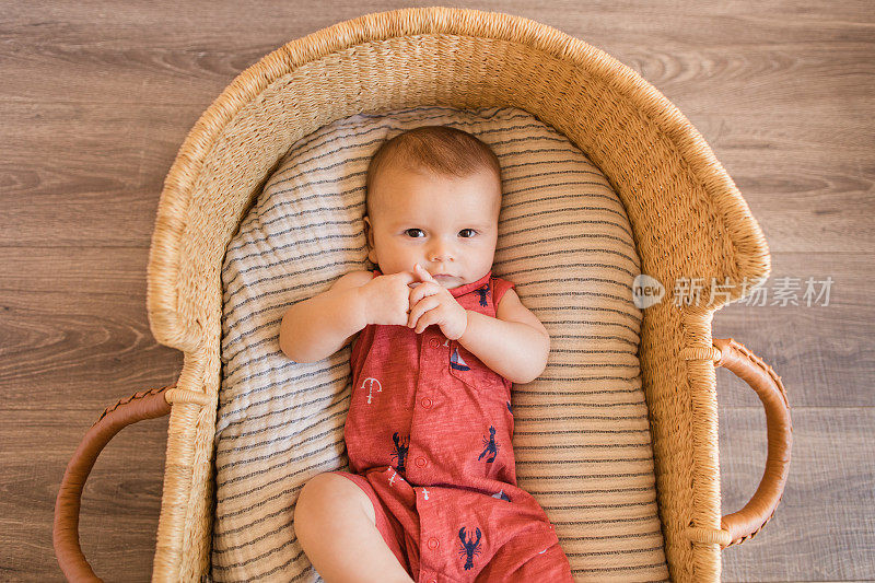 一个18周大的男婴，穿着一件红色的航海服，躺在一个舒适的奶油色条纹棉毯上，躺在一个海草摩西篮子里