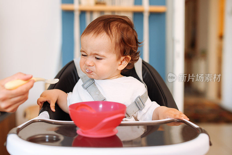 微笑的8个月大男婴坐在家里的高椅子上，妈妈用勺子给他喂固体食物