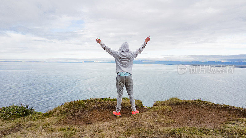 后视图十几岁的男孩举起手臂站在悬崖边，看着Jökulsárgljúfur湾的Kópasker