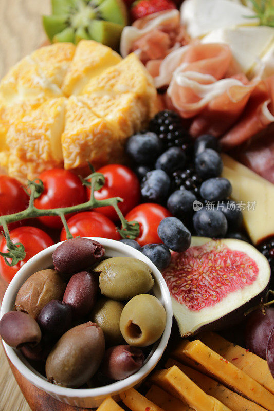 全框架图像木质熟食板的成分，布里干酪，火腿，烟熏奶酪，藤番茄，无花果，红葡萄，蓝莓，黑莓，猕猴桃，草莓，小模子的黑色和绿色橄榄，高架视图