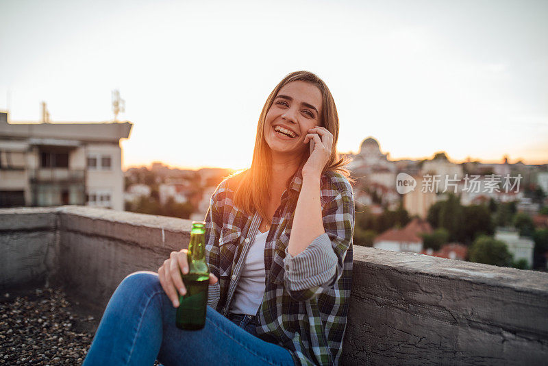 微笑的女孩喝着啤酒，在夕阳下打电话