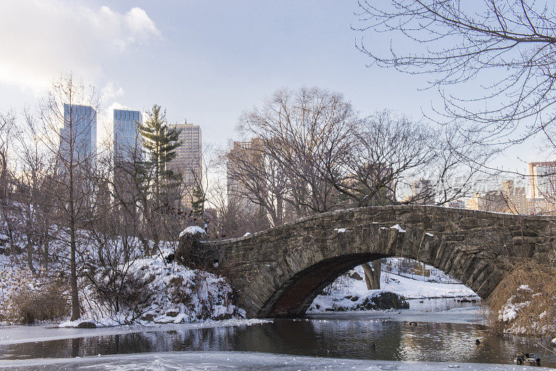 在美国纽约市曼哈顿市中心的冬天，从冰雪覆盖的中央公园，包括一个结冰的池塘和一座石桥所看到的城市景观。