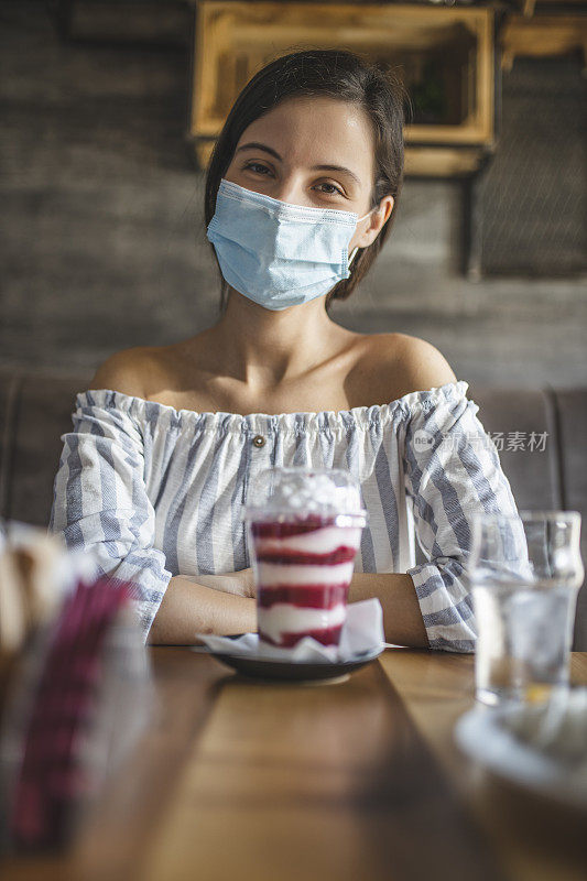 美丽的年轻女子戴着外科口罩，吃奶酪蛋糕，花时间在咖啡馆在疫情期间