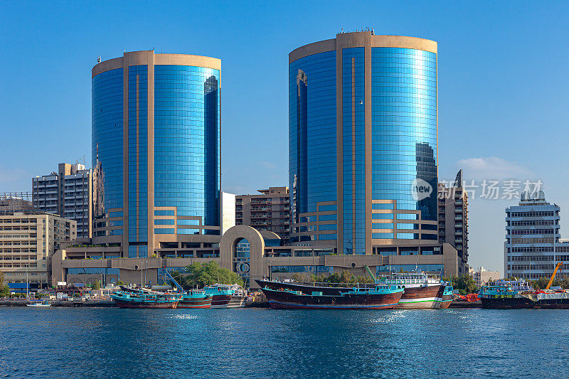 阿拉伯联合酋长国迪拜——清晨阳光下，几艘传统的阿拉伯单桅帆船并排停泊在迪拜河德拉一侧的码头上;背景中，现代办公建筑。