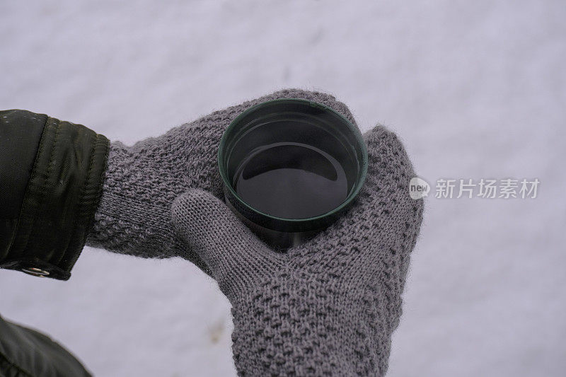 双手在温暖的针织手套握热饮料在热饮杯横跨积雪表面俯视图。冬季徒步旅行。本空间