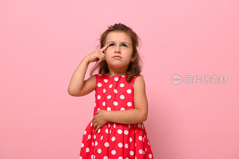 可爱可爱的学前班女孩，3-4岁，看起来若有可无，她把手指放在太阳穴上，在粉红色的背景上摆姿势。一个忧郁的小女孩的肖像