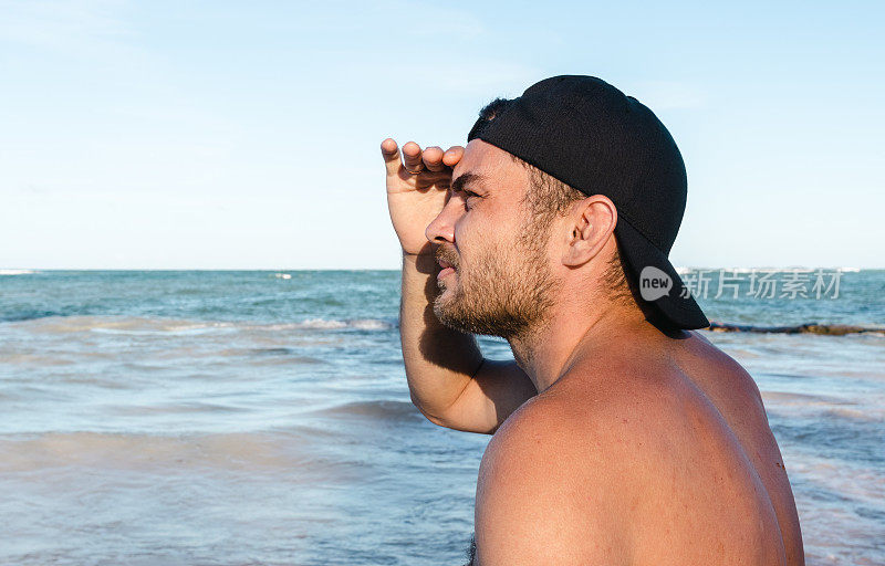 一个戴着帽子的裸着膀子的男人站在海边的沙滩上，双手举过头顶望着远方。
