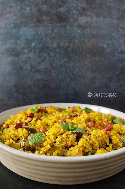 一碗印度早餐食谱poha(五香平饭)的特写图像，用洋葱，青椒，咖喱和香菜叶，花生，芥末和孜然种子，姜黄和柠檬汁，灰色背景，重点放在前景，复制空间