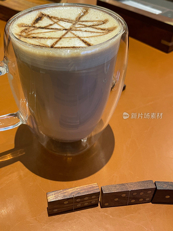 近距离图像的玻璃杯起泡咖啡，卡布奇诺咖啡杯，灰尘与巧克力粉在咖啡店的桌子上，木制多米诺骨牌瓷砖，重点在前景