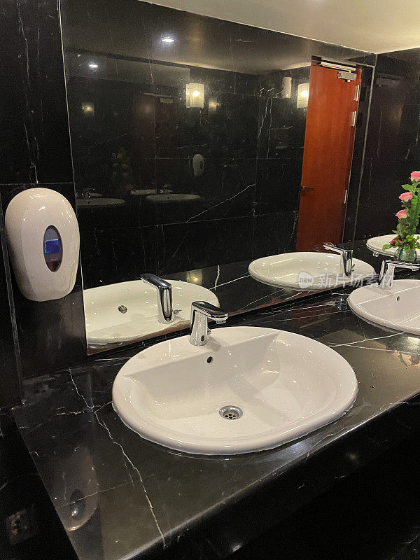 酒店公共浴室里白色的瓷水槽，金属水龙头和插头孔，黑色大理石墙壁和地板，红木门，架子上的人造玫瑰