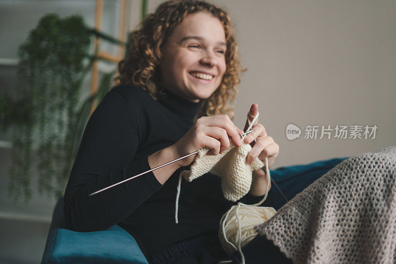 一个卷发女人坐在客厅的沙发上，为冬天织围巾。年轻女子的爱好。