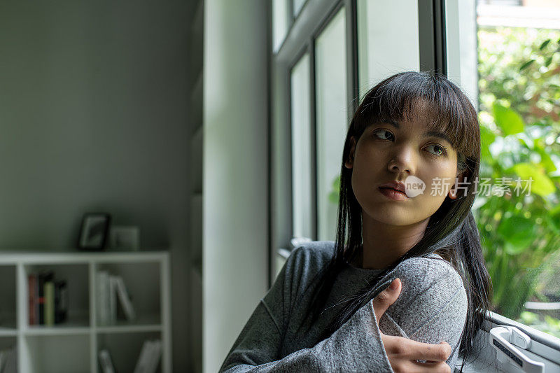 一个疲惫的年轻亚洲女子望着窗外，被压力压得喘不过气来，心理健康状况堪忧。