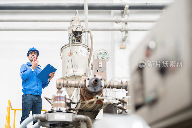 塑料和钢铁工厂的广角视图。它是由一位欧洲男性工程师管理的。使用对讲机工作靠近工作机械，手持便签，穿制服，戴安全帽，佩戴对讲机。