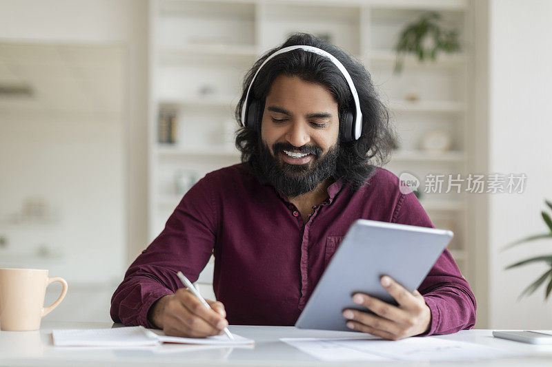 印度男子一边浏览平板电脑，一边做笔记，一边戴着耳机欣赏音乐
