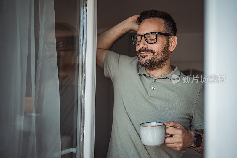 一位成年男子端着一杯咖啡，面带微笑地站在公寓的阳台门口