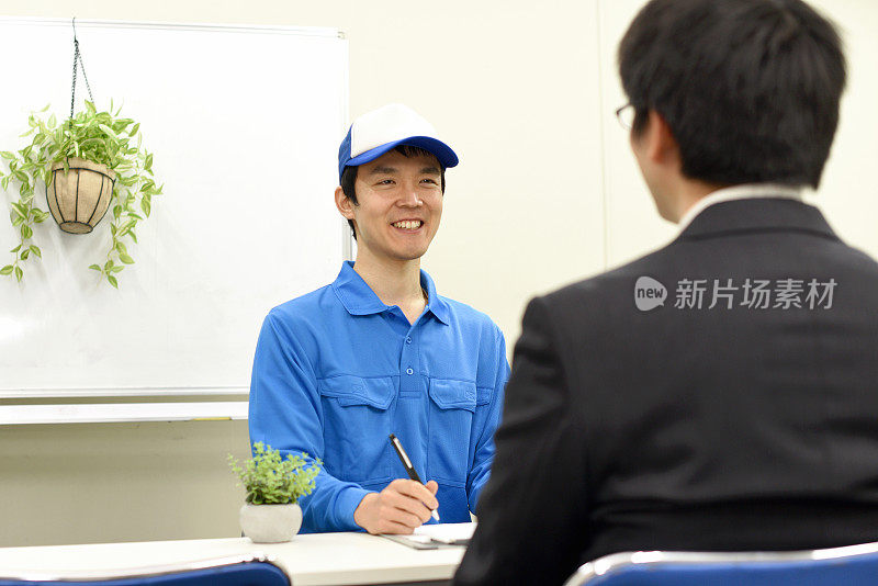 年轻的亚洲工人用着写字板，微笑着和坐在室内的亚洲男工人交谈