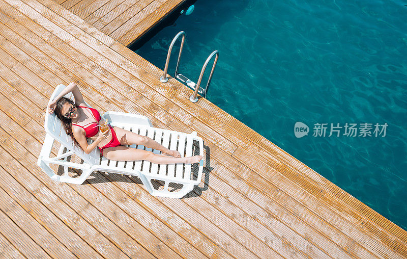 快乐的苗条女孩端着鸡尾酒躺在泳池边的躺椅上。暑期休息和假期的概念。带有复制空间的背景。