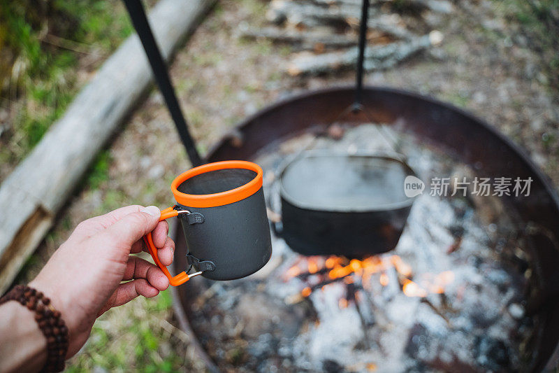 游客用马克杯沏茶，男人手里拿着杯子，背景是火，游客用器皿当食物，在大自然中露营。