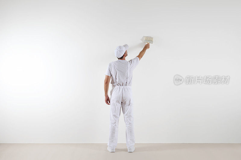 后视图画家看着和画空白的墙壁，用油漆刷，孤立在白色的大空间