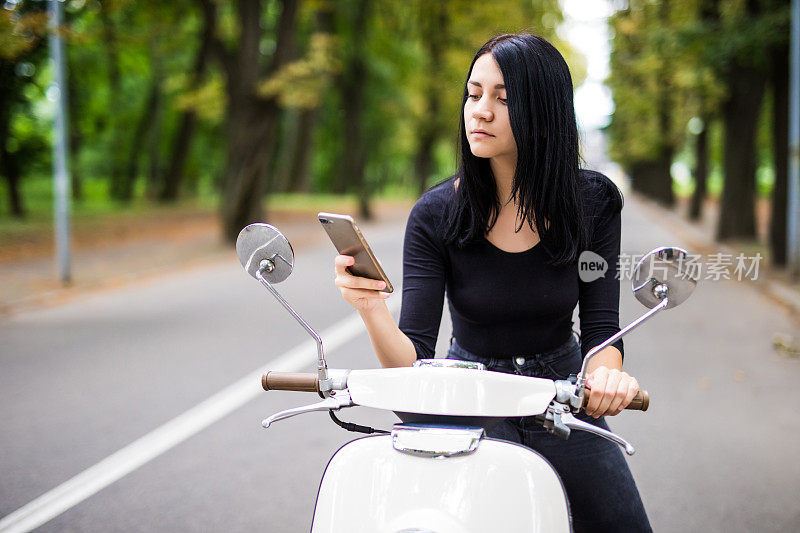 微笑的年轻女子看着她的手机，坐在摩托车上