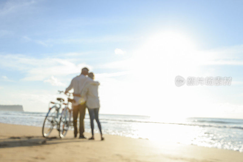 在日落或日出时在海滩上骑自行车的成熟夫妇的散焦图像。