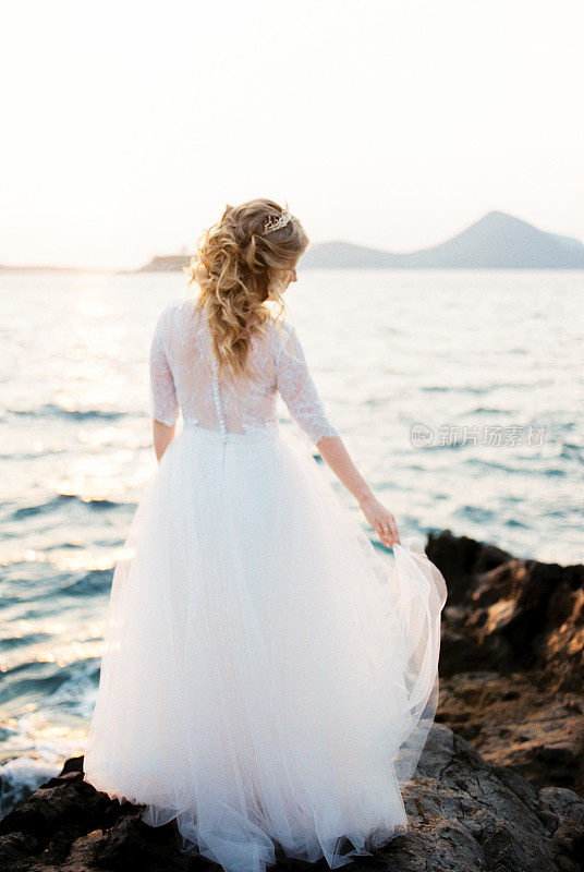 新娘在海边的岩石上拿着一件层层叠叠的连衣裙的下摆。后视图