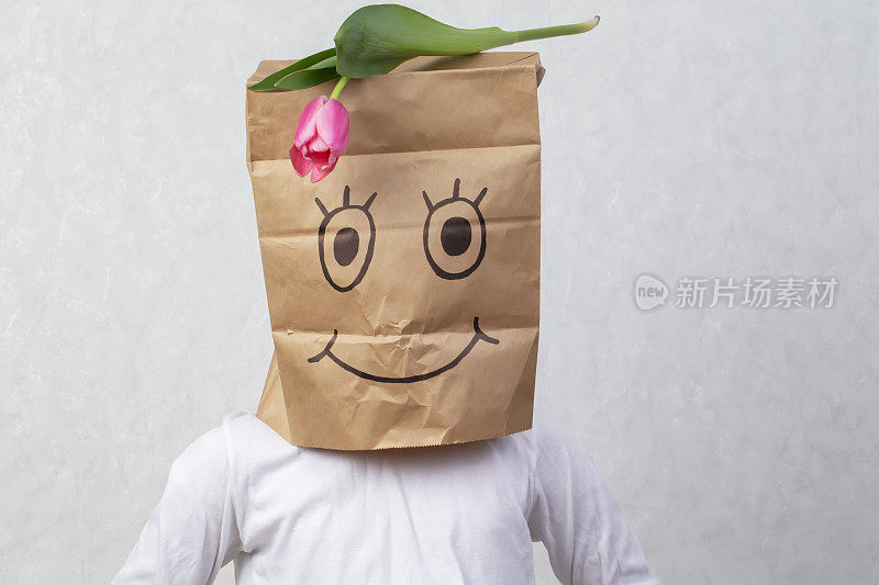 一个有趣的男孩把一个画着微笑的纸袋放在他的头上，并在上面放了一朵郁金香。匿名礼物的概念。