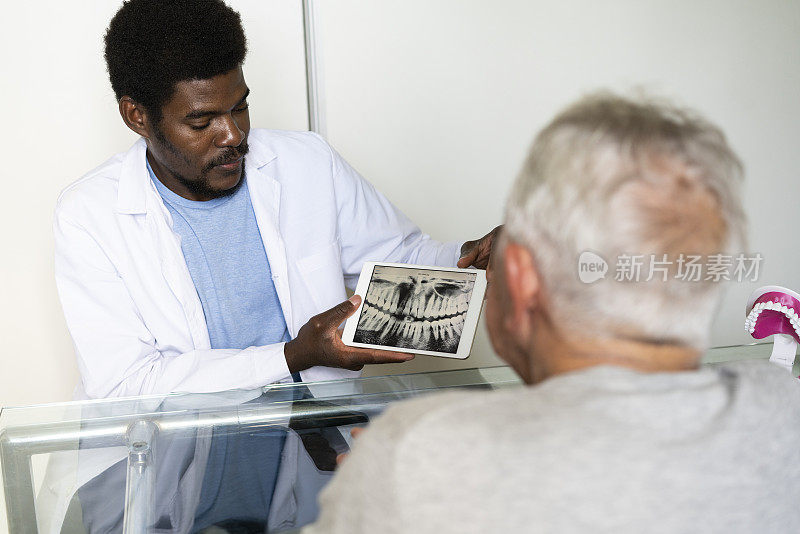 非裔美国男性牙医在她的办公室里通过电子平板电脑向老年病人展示全景颌x光