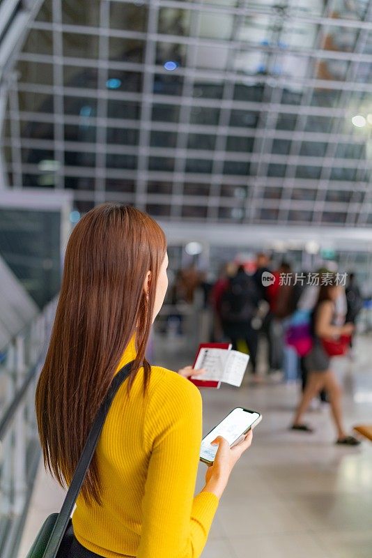 年轻的亚洲女性在机场出发大厅，拿着智能手机、护照和登机牌，一边听音乐一边等待登机