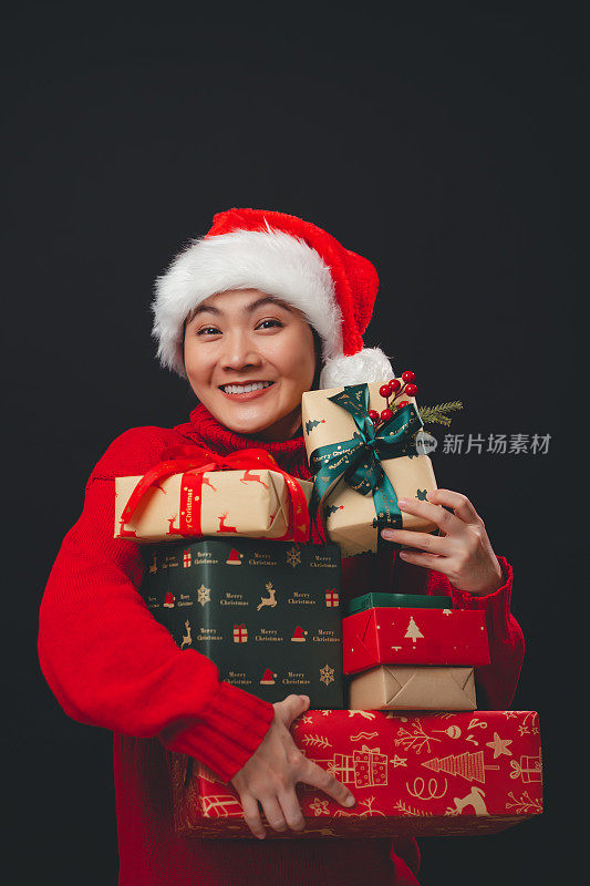 亚洲女人戴着圣诞老人的帽子开心地微笑着拿着许多礼盒，孤立在黑色的背景上。