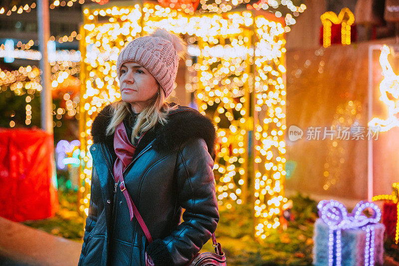 穿着冬季夹克的妇女站在装饰着彩灯的圣诞市场旁，望着别处