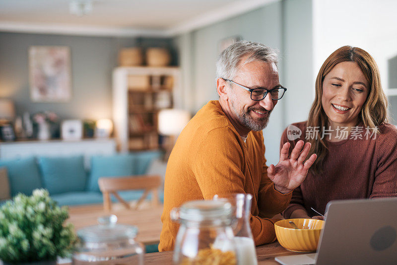 幸福的成熟夫妇在家里用笔记本电脑视频通话时向某人挥手。