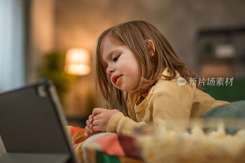 一个蹒跚学步的女孩躺在地板上用平板电脑看动画片，吃着爆米花