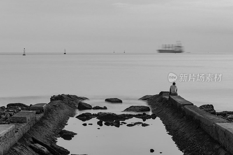 一个孤独的人坐在水渠的墙边看海。下午晚些时候海滩上的时间流逝。一艘货船在地平线上驶过。巴西桑托斯市。黑白图像。