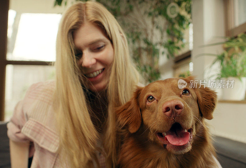 一位女士和心爱的宠物狗新斯科舍省鸭鸣猎犬一起躺在沙发上，愉快地抚摸着它