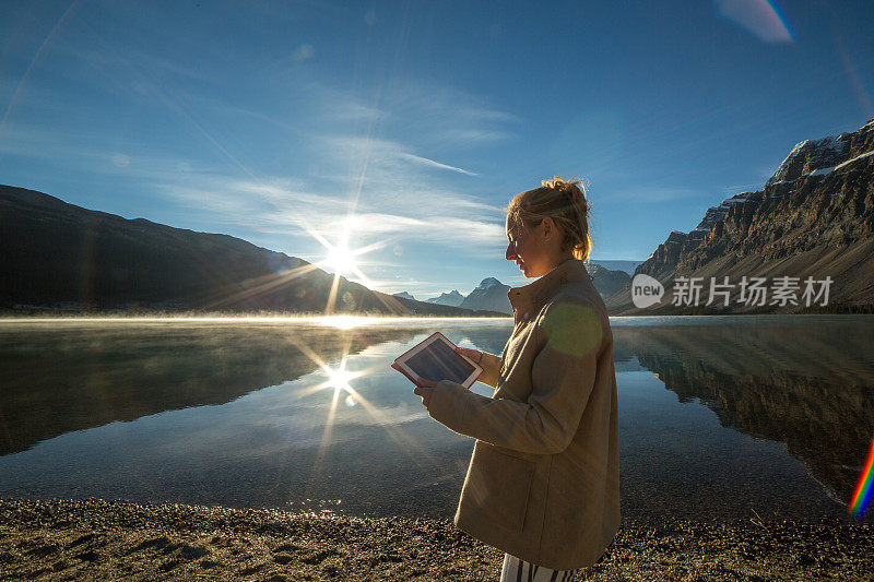一名女子走在湖边，用数码平板电脑拍摄风景