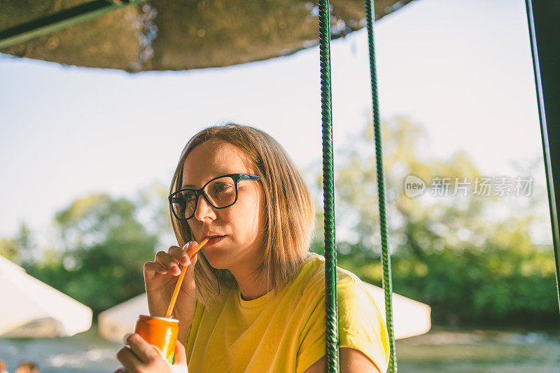 年轻美丽的海滩女孩。侧面的美丽和年轻的女人在泳装和太阳镜保持玻璃与橙汁在手和微笑而坐在木制码头。