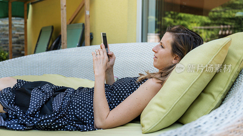 年轻女子躺在露台沙发上浏览她的手机
