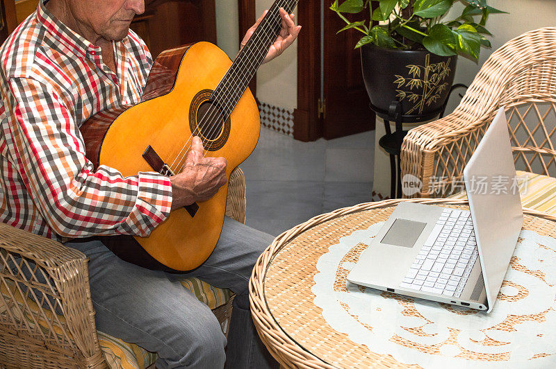 一位老人在家里用电脑学习弹吉他。老人与技术，远程课程或在线教程的概念。