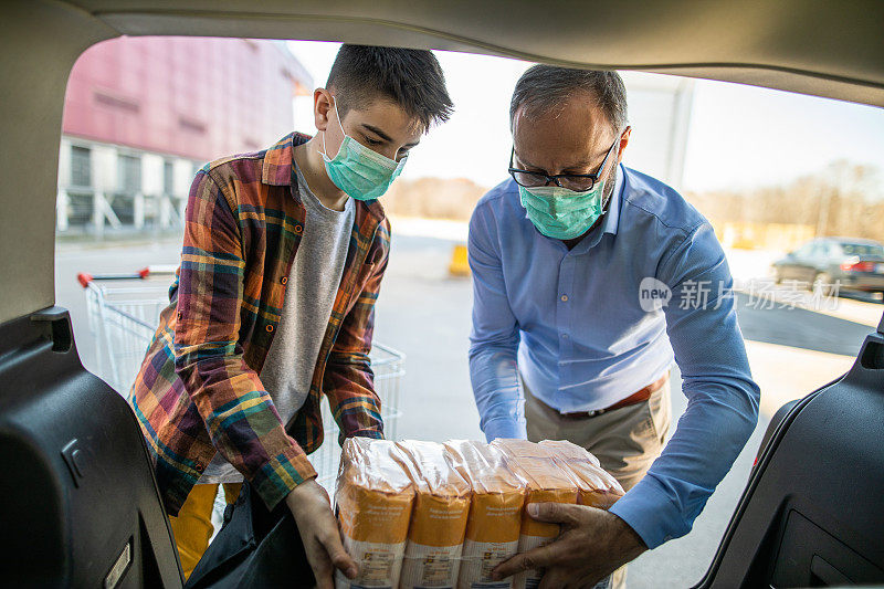 在病毒大流行期间，父亲和儿子戴着防护口罩往汽车后备箱装食物