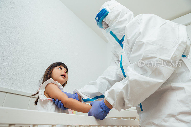 身穿个人防护装备的医生在家中的婴儿床上照料一位微笑着的黑头发COVID-19患者小女孩