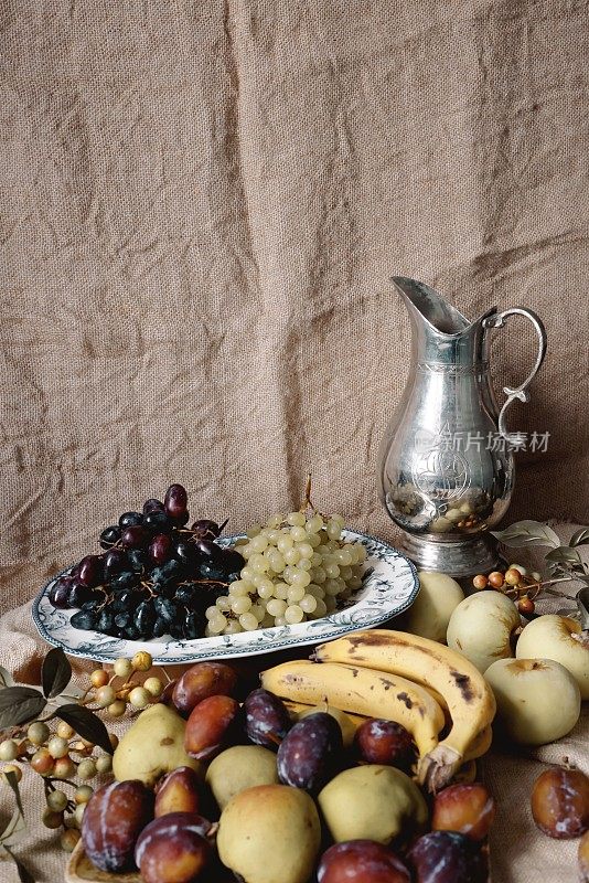 一个经典的银壶，周围是冬天的浆果，背景是粗麻布，盘子里有红的和黄的葡萄，西洋李子，青苹果，香蕉，梨