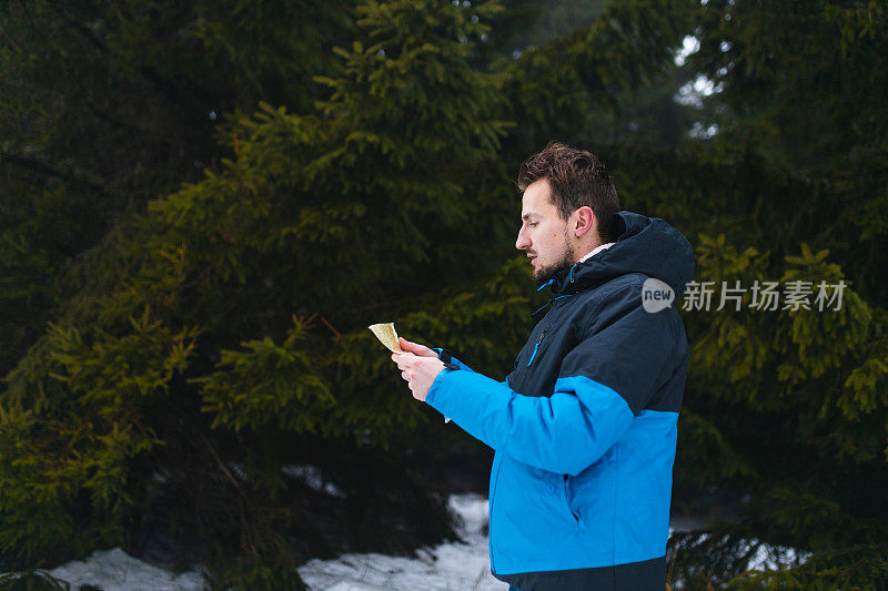 年轻的男性徒步旅行者穿着蓝色夹克，在雾蒙蒙的雪山中迷失了方向，看着地图