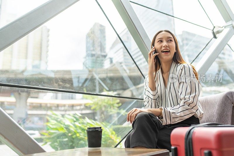 美丽的年轻亚洲女性商务独自女旅行者在机场候机楼的咖啡馆里等待起飞时用智能手机打电话