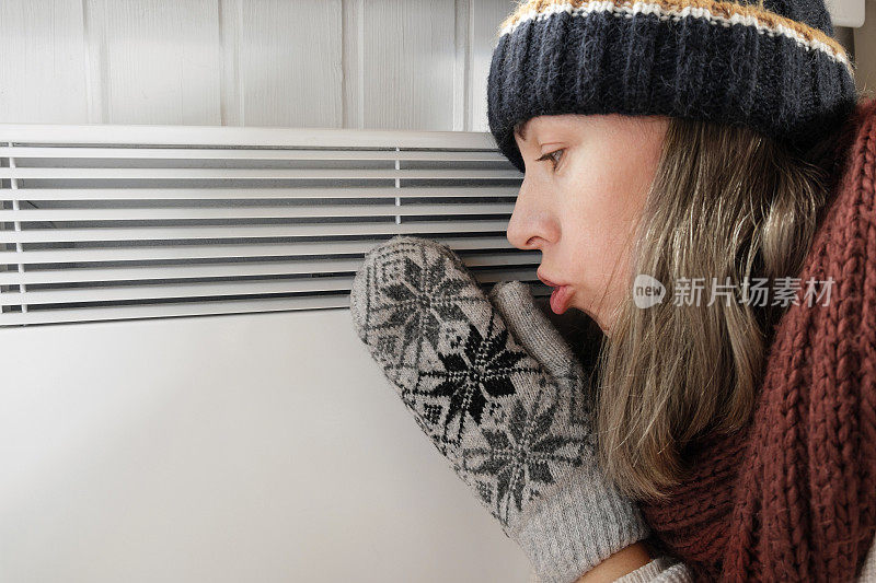 年轻的冷冻妇女穿着毛衣颤抖和冻结的冬天寒冷与震惊的表情在脸上。女孩正在电暖炉上用呼吸暖手。不舒服的呆在家里。